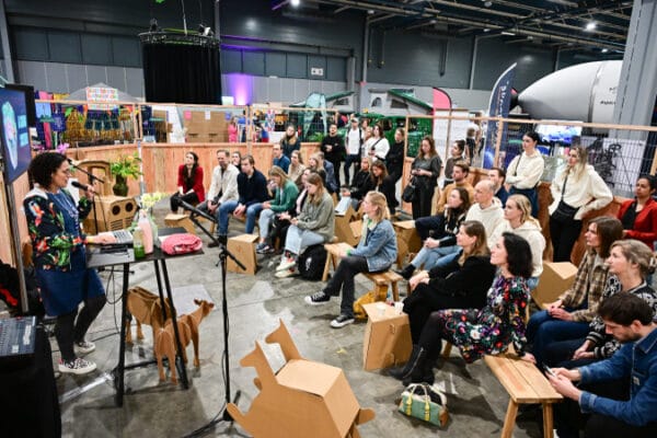 Een spreker geeft een presentatie aan een aandachtig publiek tijdens de Impact Fair 2025. De presentatie vindt plaats in een informeel ingerichte ruimte met kartonnen zitplaatsen en decoratieve elementen. Op de achtergrond is de SpaceBuzz-raket zichtbaar.
