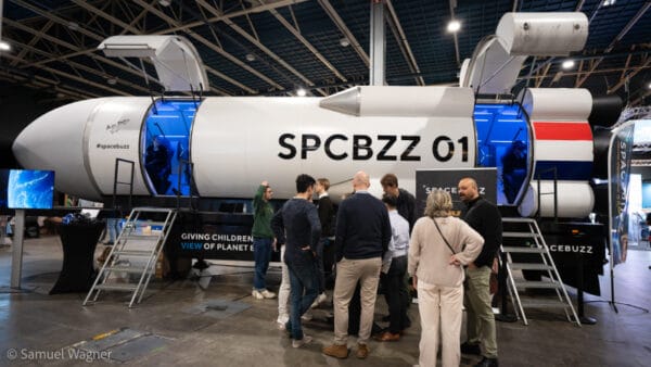 De SpaceBuzz-raket bij de Impact Fair 2025. Bezoekers staan rondom een futuristische VR-raket, waarbinnen kinderen een virtual reality-ervaring beleven over de aarde vanuit de ruimte. Op de raket staat "SPCBZZ 01" en een scherm toont een beeld van de aarde vanuit de ruimte.