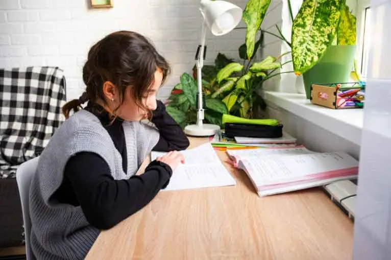 Kind oefent met opdrachten uit een oefenboek aan een bureau bij het raam.