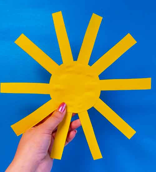 Hand houdt een gele papieren zon met geknipte zonnestralen tegen een blauwe achtergrond.